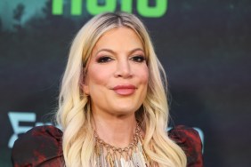 Tori Spelling smiling in front of a black back drop, wearing a gold necklace and red jacket