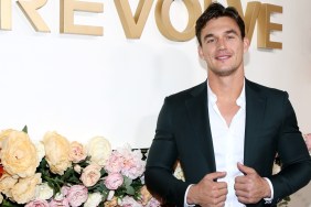 Tyler Cameron posing next to a flower arrangement, his hands are on the jacket of his open suit and he isn't wearing a tie