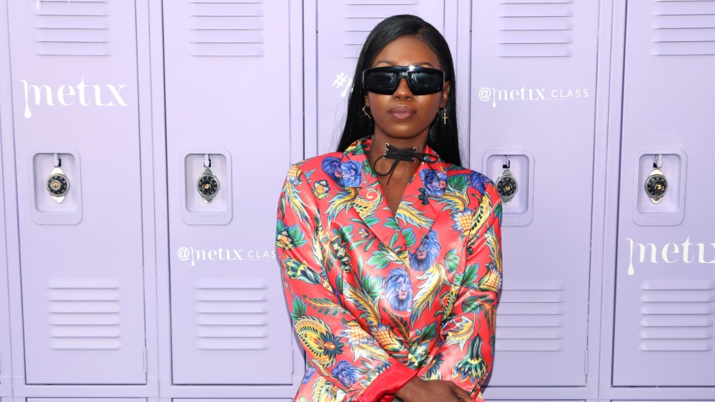 Faith Stowers posing in front of purple lockers, she's wearing a multicolored blazer and has sunglasses on.