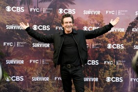 Jeff Probst in an all-black outfit holding his hands up in front of a Survivor backdrop