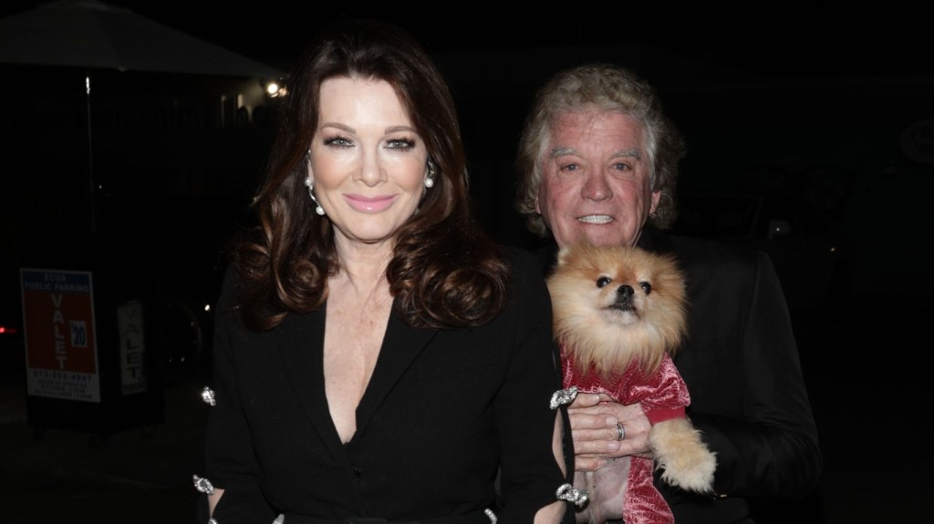 Lisa Vanderpump and Ken Todd wearing all black and holding up a dog wearing a red sweater