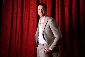 Dylan Efron in a grey suit standing in front of a red curtain